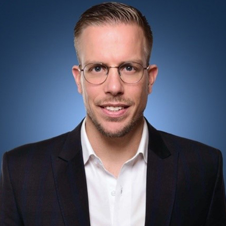 Persoon met bril draagt een blazer en overhemd tegen een blauwe achtergrond.