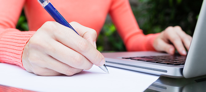 Une femme, assise dans un jardin, écrit à la main une lettre de démission tout en consultant des informations sur son ordinateur portable.