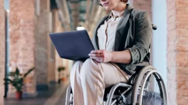 Person using a laptop while sitting in a wheelchair in a bright hallway.