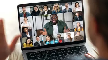 Person waving at a laptop screen during a video call with multiple participants smiling.