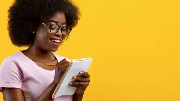 Person holding a notebook and pencil, smiling against a yellow background