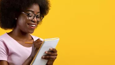 Person writing in a notebook, smiling against a yellow background.