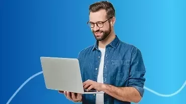 A man wearing glasses, smiling and standing, holding a laptop aloft. He looks happy and excited.