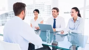 Person seated at a table with three others, all dressed in business attire, engaged in a meeting in a bright office.