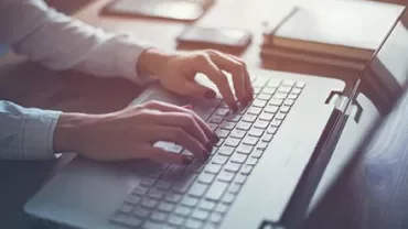Person typing on a laptop at a desk, with notebooks and a pen nearby.