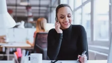 Persoon in coltrui die aan de telefoon praat en schrijft aan een bureau in een modern kantoor.
