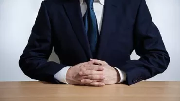 Person in a suit sitting at a wooden table with hands clasped.
