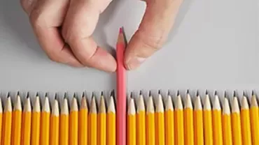 Hand holding a pink pencil among yellow pencils on a grey surface.