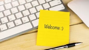 Yellow sticky note with 'Welcome :)' on a desk beside a keyboard and pen.