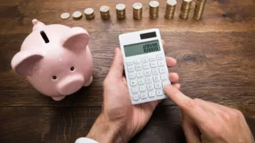 Person using a calculator next to a piggy bank and stacked coins on a wooden table.