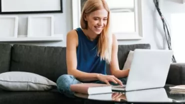 Persoon zit op een bank en gebruikt een laptop, glimlachend.