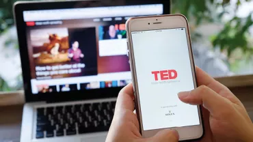 Persoon houdt telefoon met TED-app geopend, laptop op de achtergrond.