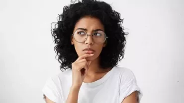 Personne aux cheveux bouclés et lunettes pensant, main sur le menton.