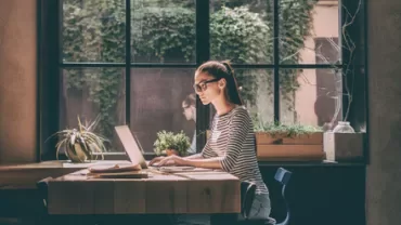 Personne travaillant sur un ordinateur portable à une table ensoleillée avec des plantes et une grande fenêtre.