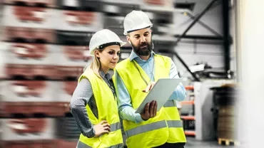 Two workers in safety vests and helmets discuss over a tablet in a warehouse.