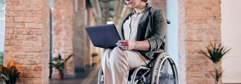 Person using a laptop while sitting in a wheelchair in a bright hallway.