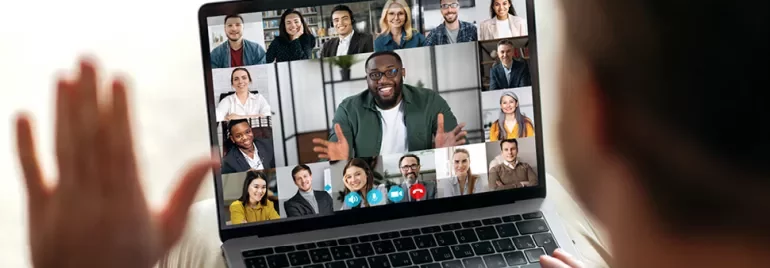 Personne saluant un écran d'ordinateur portable lors d'un appel vidéo avec de nombreux participants souriants.