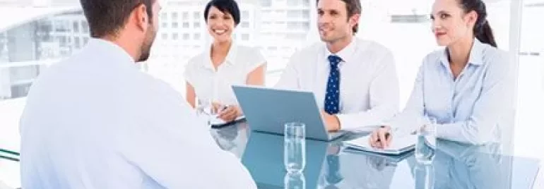 Personne assise à une table avec trois autres, tous en tenue de bureau, participent à une réunion dans un bureau lumineux.