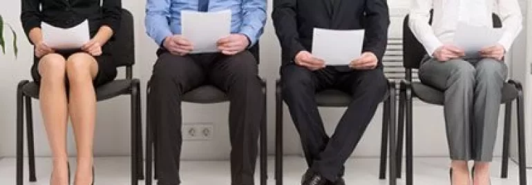 Four people sitting in chairs holding papers, dressed in business attire.