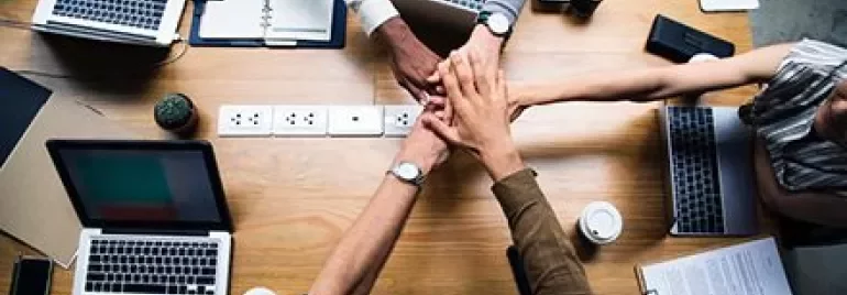 Groupe de personnes joignant les mains au-dessus d'un bureau avec des ordinateurs portables et du café.
