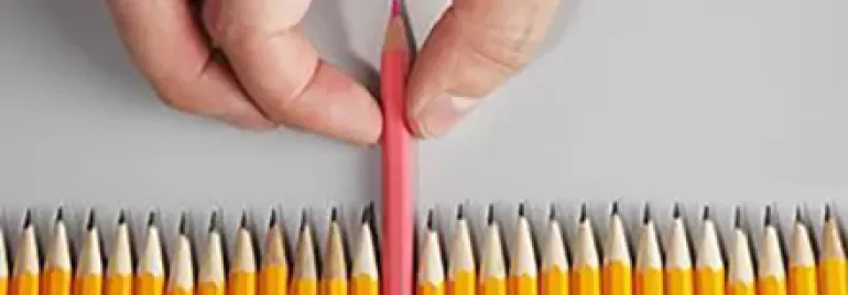 Main tenant un crayon rose parmi des crayons jaunes sur une surface grise.