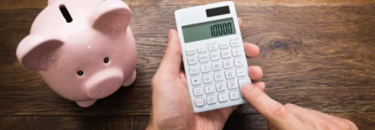 Personne utilisant une calculatrice à côté d'une tirelire et de pièces empilées sur une table en bois.
