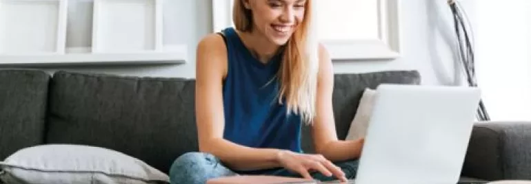 Persoon zit op een bank en gebruikt een laptop, glimlachend.