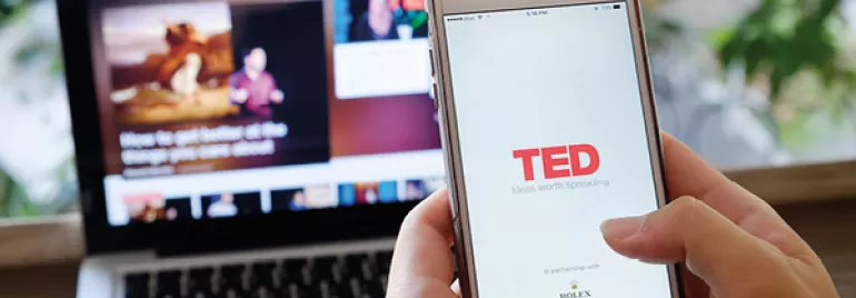 Persoon houdt telefoon met TED-app geopend, laptop op de achtergrond.