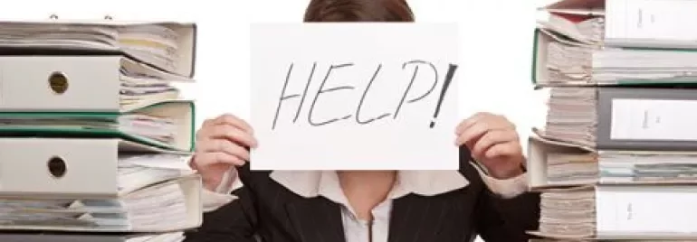Person holding 'HELP!' sign behind stacks of folders.