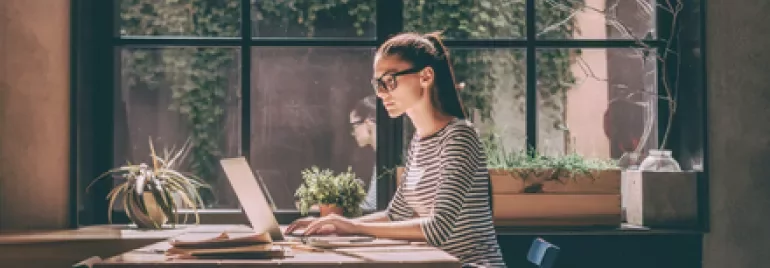 Person working on a laptop at a sunlit table with plants and a large window.