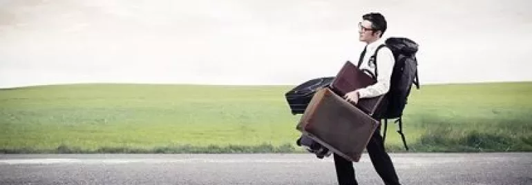 Person walking on a road with suitcases and a backpack in open countryside.