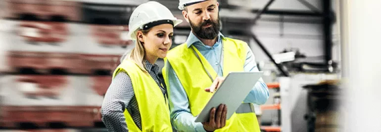 Two workers in safety vests and helmets discuss over a tablet in a warehouse.