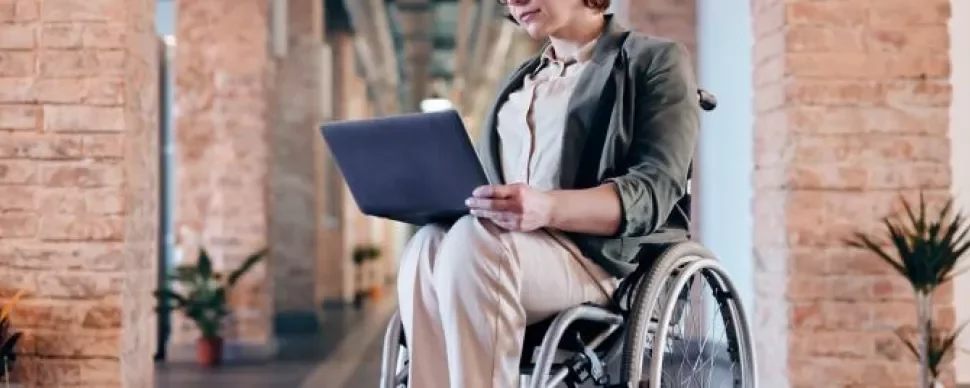 Person using a laptop while sitting in a wheelchair in a bright hallway.