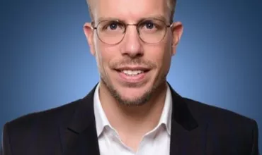 Person with glasses wearing a blazer and shirt against a blue background.