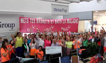 Group of people in an office holding a large check, wearing colorful clothes, with a pink banner in the background.