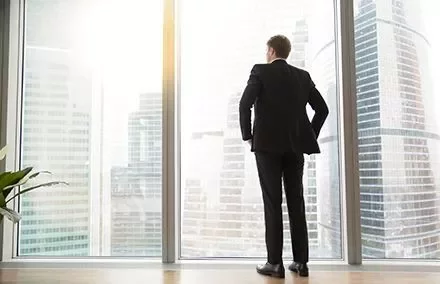 Person in suit looks out at cityscape through tall windows.