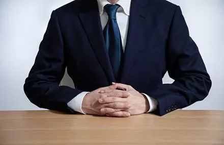 Person in a suit sitting at a wooden table with hands clasped.