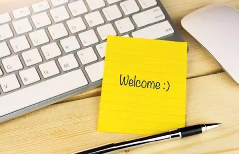 Gele plaknotitie met 'Welcome :)' op een bureau naast een toetsenbord en pen.