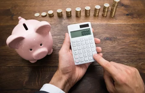 Person using a calculator next to a piggy bank and stacked coins on a wooden table.