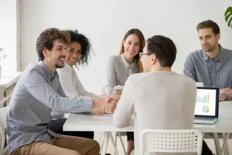 Mensen zitten rond een tafel tijdens een vergadering, één persoon schudt de hand van een andere.