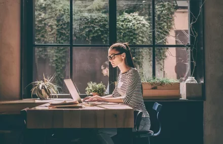 Persoon werkt op een laptop aan een zonnige tafel met planten en een groot raam.