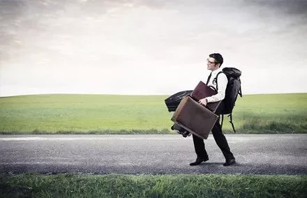 Person walking on a road with suitcases and a backpack in open countryside.